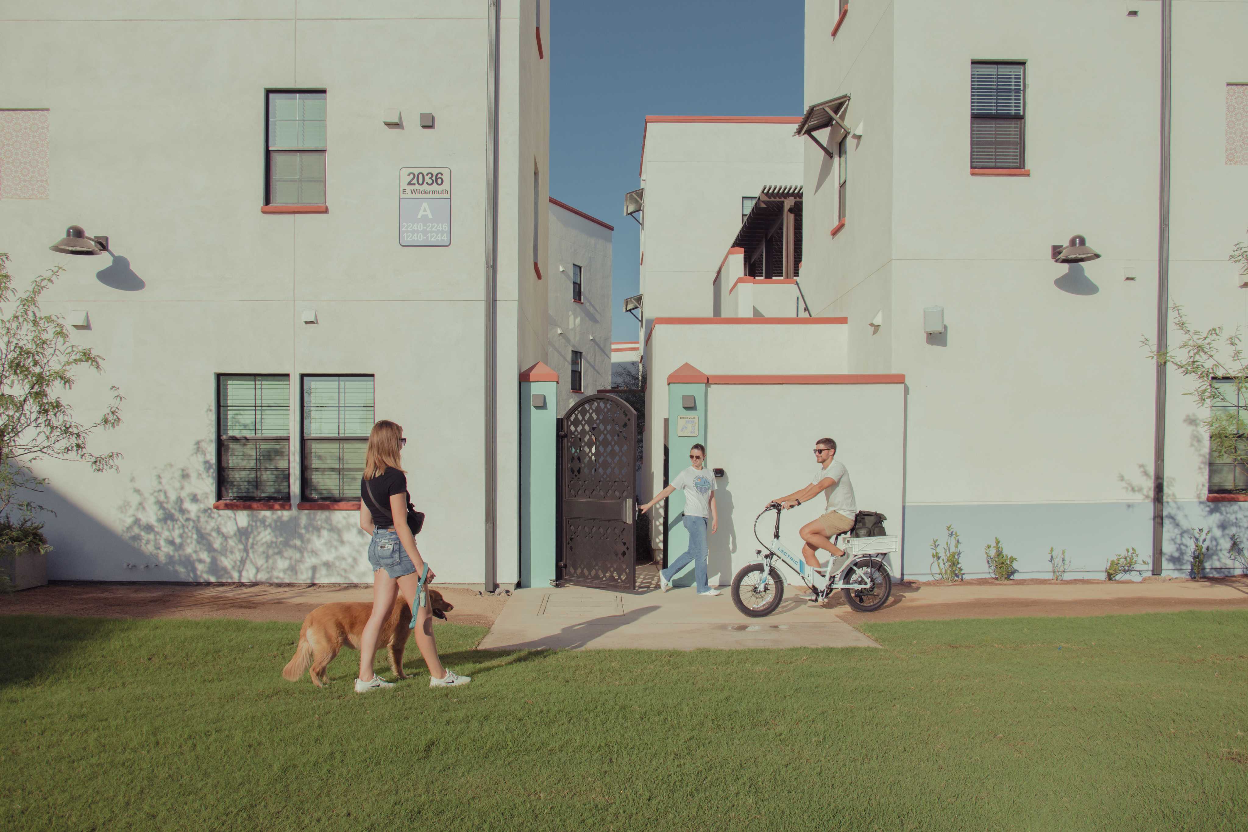 Woman walking dog talking to another woman exiting a neighborhood area and a man on an ebike